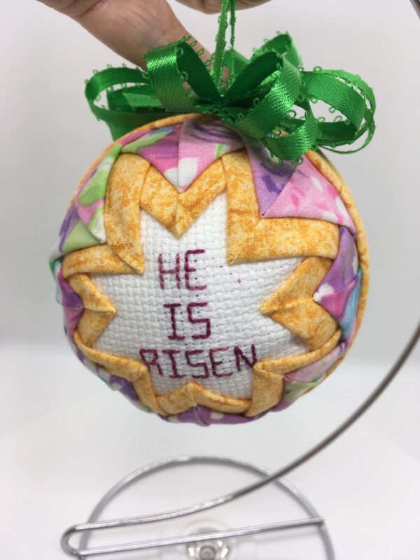 A three inch ornament has the cross stitch message, He is Risen, in the center, frame by golden yellow fabric folded and pinned to a polystyrene ball. The yellow is framed by a rose pastel fabric.