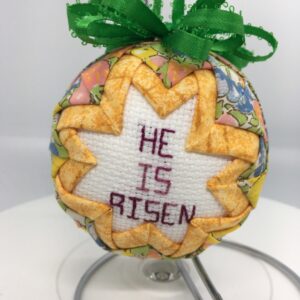 A cross-stitch message of He Is Risen is surround by golden yellow fabric pinned on a three inch polystyrene ball, then surrounded by pastel fabric.