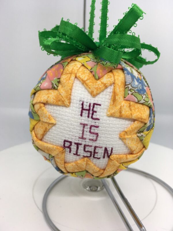 A cross-stitch message of He Is Risen is surround by golden yellow fabric pinned on a three inch polystyrene ball, then surrounded by pastel fabric.
