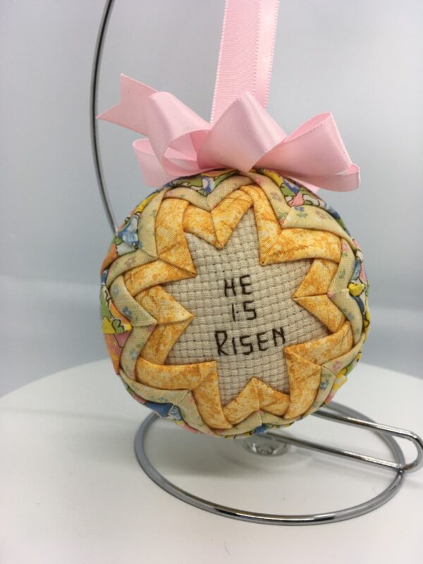 A three inch ornament has the cross stitch message, He is Risen, in the center, frame by golden yellow fabric folded and pinned to a polystyrene ball. The yellow is framed by a pale pastel fabric.
