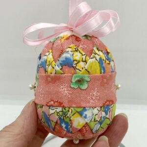 Pale orange and a yellow prints fabrics are folded and pinned on a three inch egg shape foam. It is topped with a pale pink row and ribbon.
