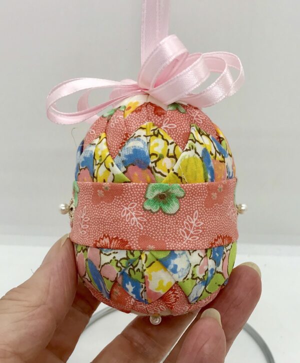 Pale orange and a yellow prints fabrics are folded and pinned on a three inch egg shape foam. It is topped with a pale pink row and ribbon.