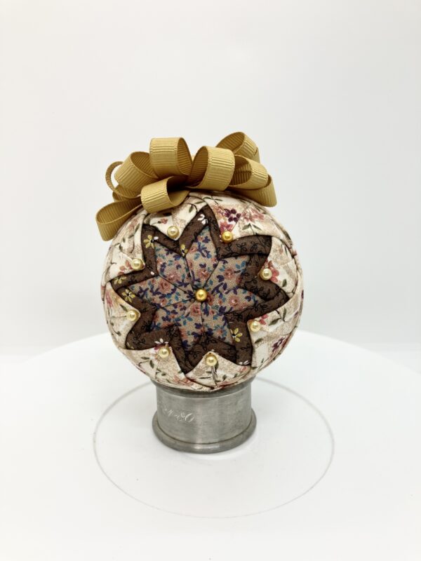 Three inch round ornament with folded fabric to create a basic star using brown calico fabric in the center, framed with darker brown fabric and then a cream calico. It is topped with gold ribbon bow and hanger.