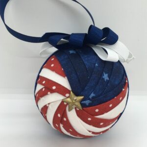 3 inch round ornament made from folded red, white and blue fabrics, in a pattern called Sparkler. Red fabric has white polka dots and blue fabric has stars. It is topped with a blue blow and blue hanger.