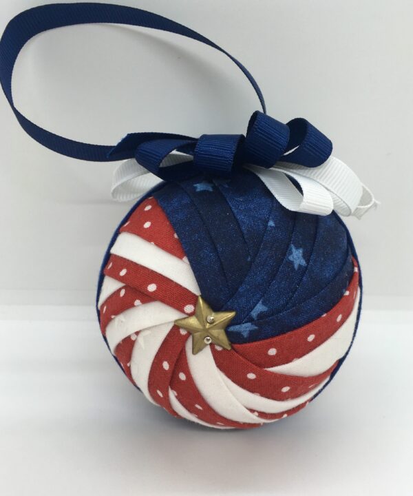 3 inch round ornament made from folded red, white and blue fabrics, in a pattern called Sparkler. Red fabric has white polka dots and blue fabric has stars. It is topped with a blue blow and blue hanger.