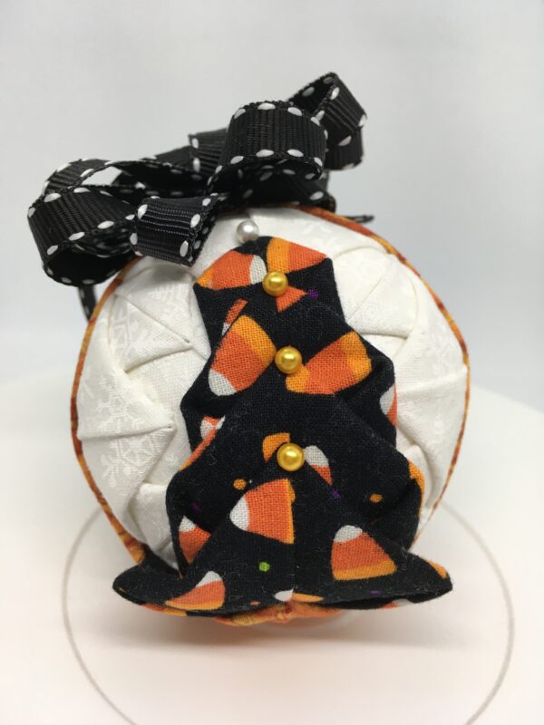 Three inch round ornament made with folding and pinning fabrics on the a polystyeren ball. The candy corn fabric with black background are folded to resemble a tree. White fabric makes the backgroun and a pumpkin print is used as a band to separate one side from the other. It it topped with a black ribbon with white edging as a bow and hanger.