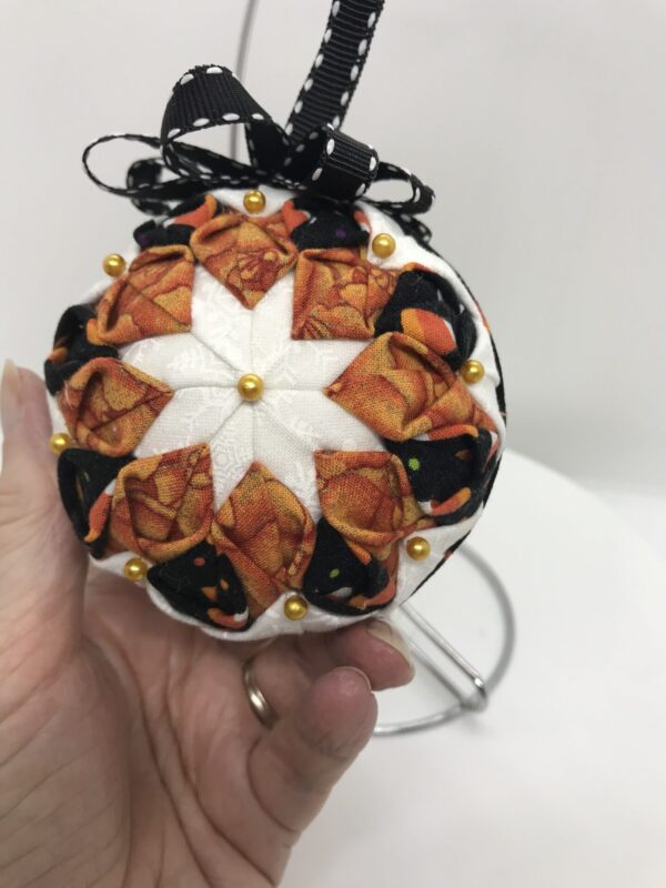 Three inch round ornament with a two rows of folded fabric to create a double bloom. Fabric colors include pumpkin print, candy corn print and white. It's topped with a black ribbon bow edged in white.