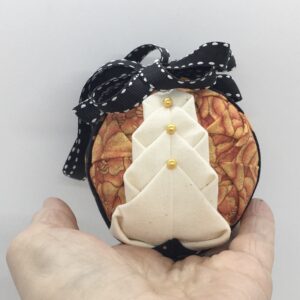 Three inch round ornament made with folding and pinning fabrics on the a polystyeren ball. Winter white fabric is folded to resemble a tree and is pinned on a background of pumpkin print fabric. It it topped with a black ribbon with white edging as a bow and hanger.