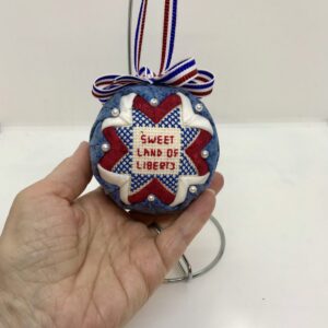 A hand is holding a three inch patriotic ornament which has the words, Sweet Land of Libery, cross stitched in the center. It is framed with red, white and blue fabrics. A bow and hanger are attached, which are also red, white and blue.