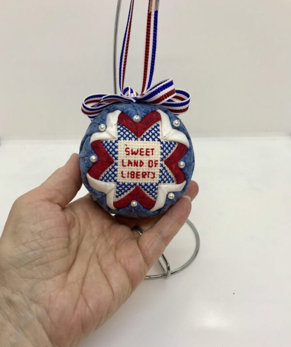 A hand is holding a three inch patriotic ornament which has the words, Sweet Land of Libery, cross stitched in the center. It is framed with red, white and blue fabrics. A bow and hanger are attached, which are also red, white and blue.