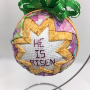 A three inch ornament has the cross stitch message, He is Risen, in the center, frame by golden yellow fabric folded and pinned to a polystyrene ball. The yellow is framed by a rose pastel fabric.
