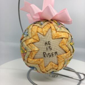 A three inch ornament has the cross stitch message, He is Risen, in the center, frame by golden yellow fabric folded and pinned to a polystyrene ball. The yellow is framed by a pale pastel fabric.