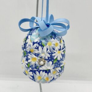 Blue daisy fabric and other blue fabrics are folded and pinned onto a three inch egg-shaped foam. It is topped with blue bown and ribbon.