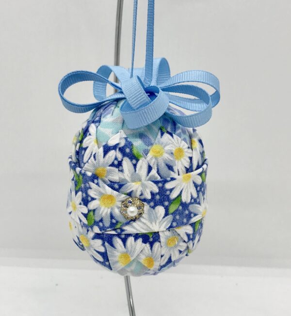 Blue daisy fabric and other blue fabrics are folded and pinned onto a three inch egg-shaped foam. It is topped with blue bown and ribbon.